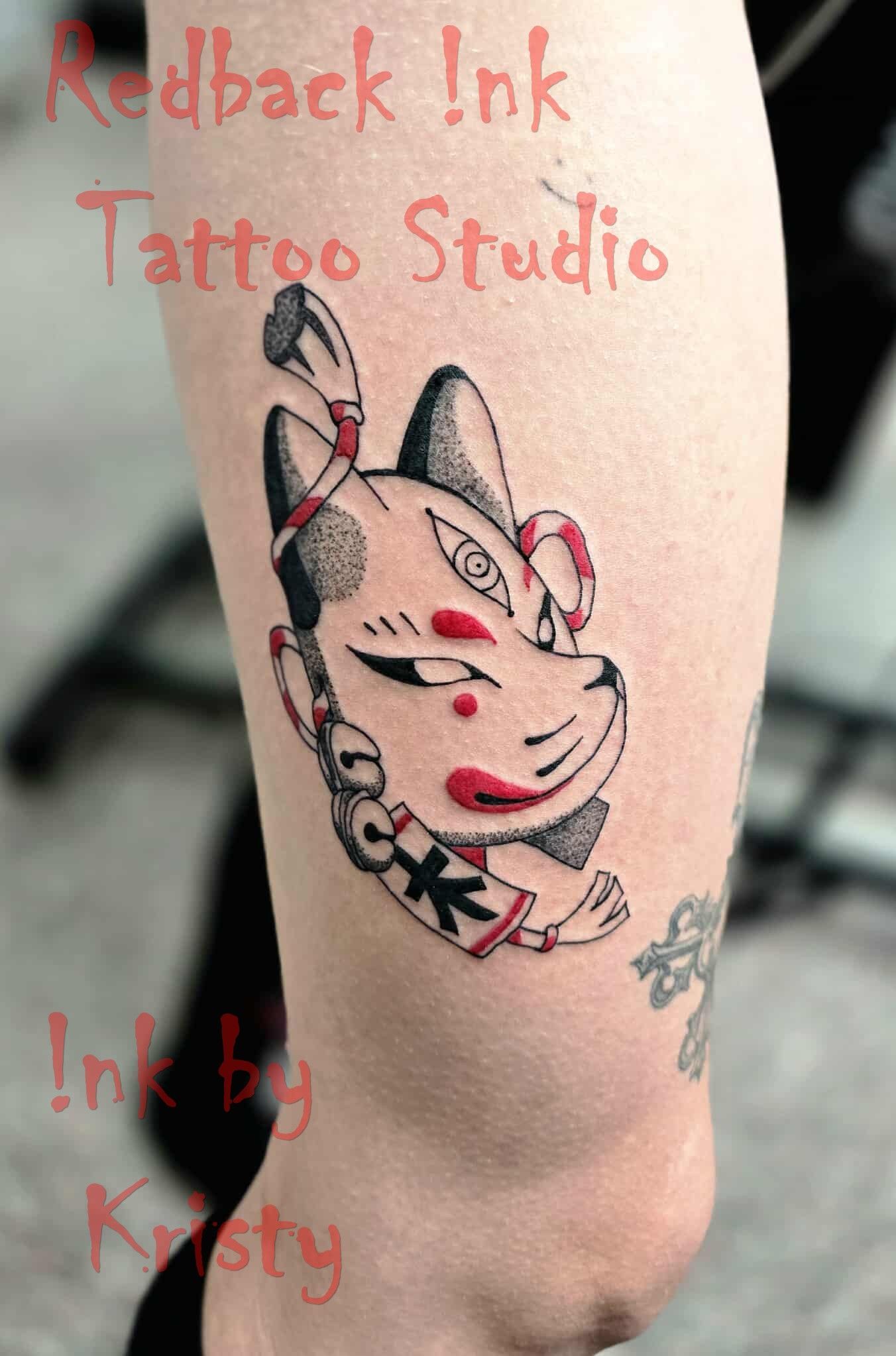 Tattoo of a Kitsune mask with red accents on a leg. Text: 