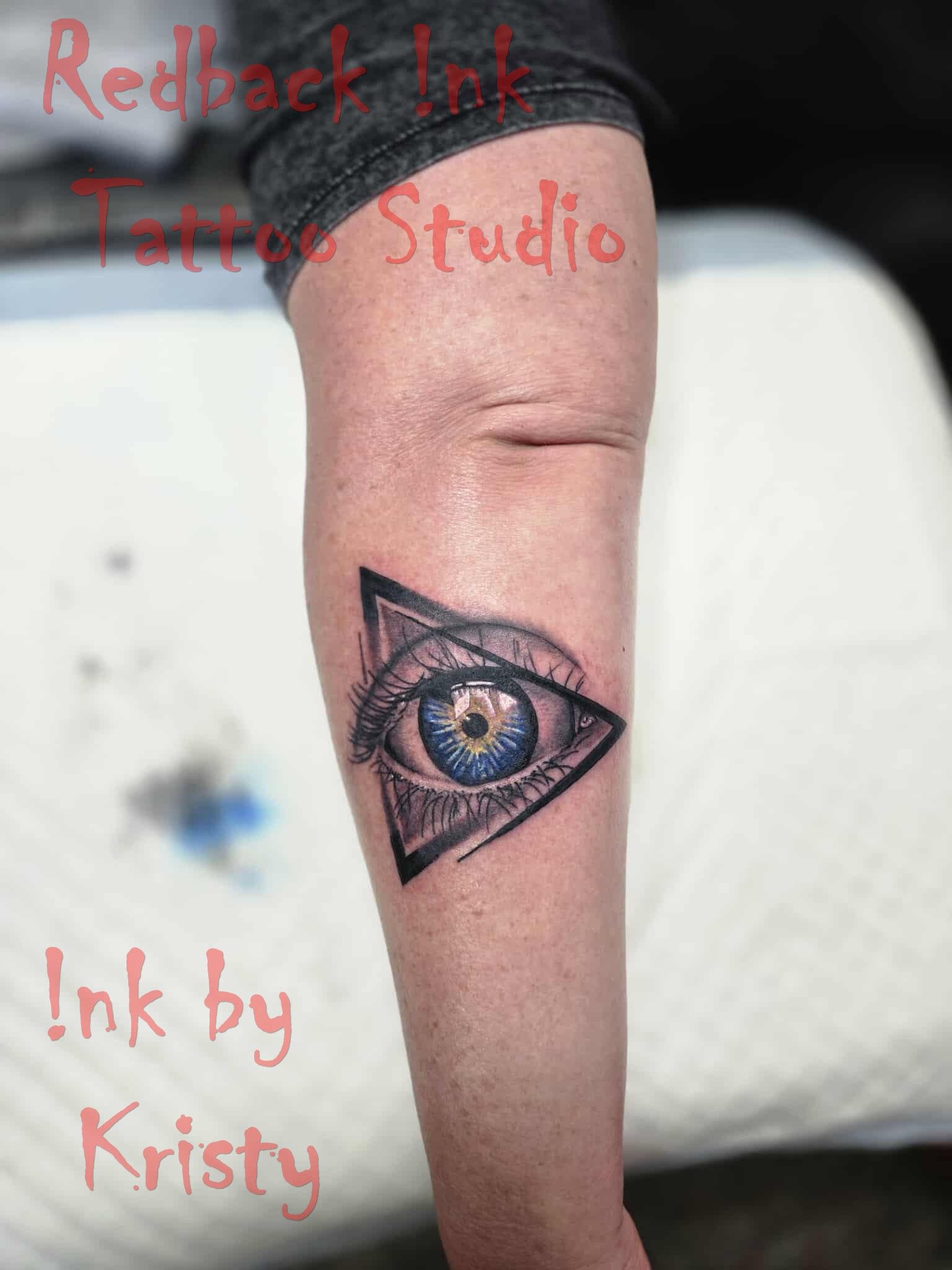 Tattoo of a blue-eyed eye within a triangle on a person's forearm, with 