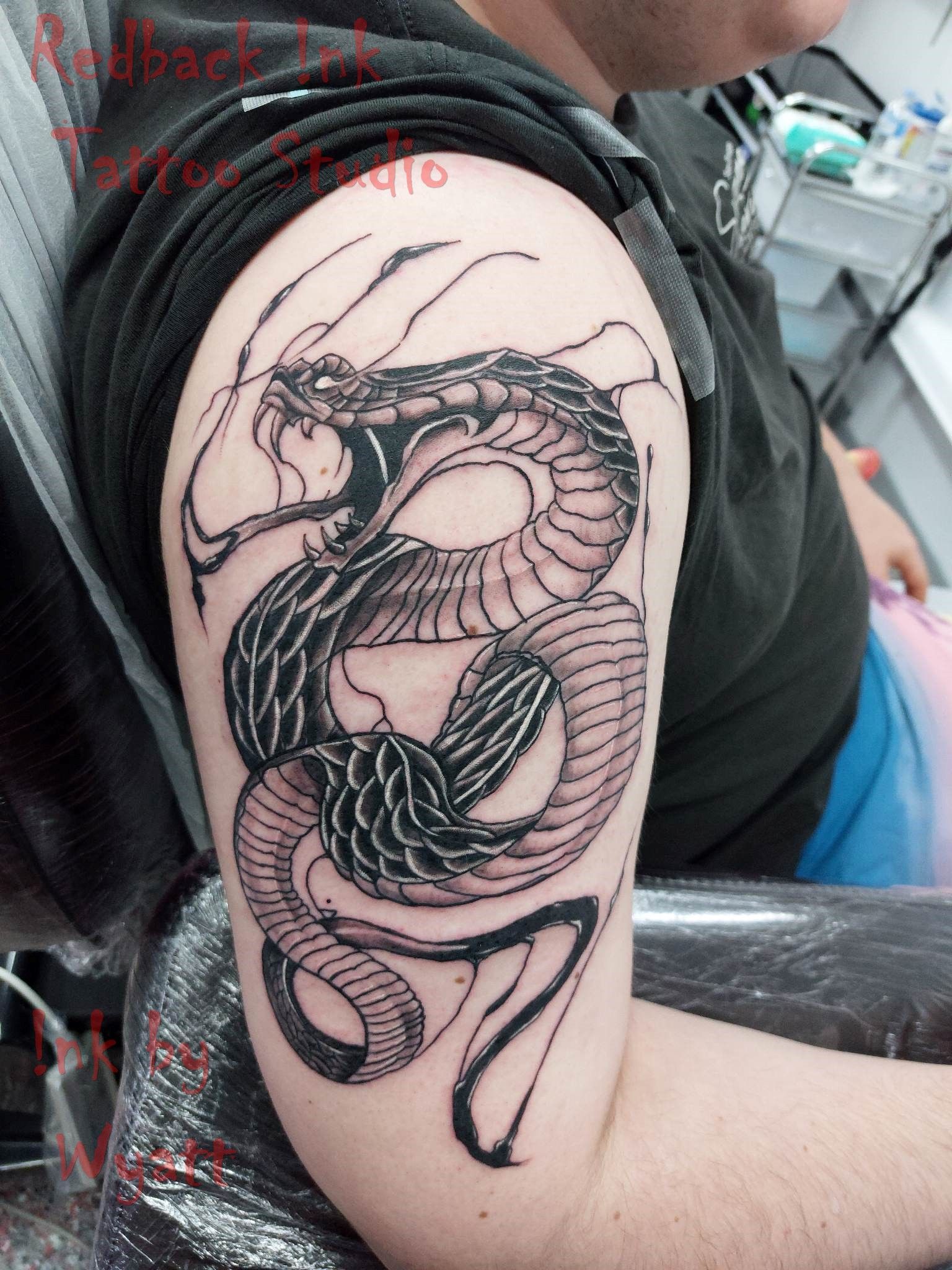 Black ink tattoo of a cobra coiled on a person's upper arm; the cobra is stylized, with detailed scales and an open mouth. — Redback !nk Tattoo Studio in Kawana, QLD