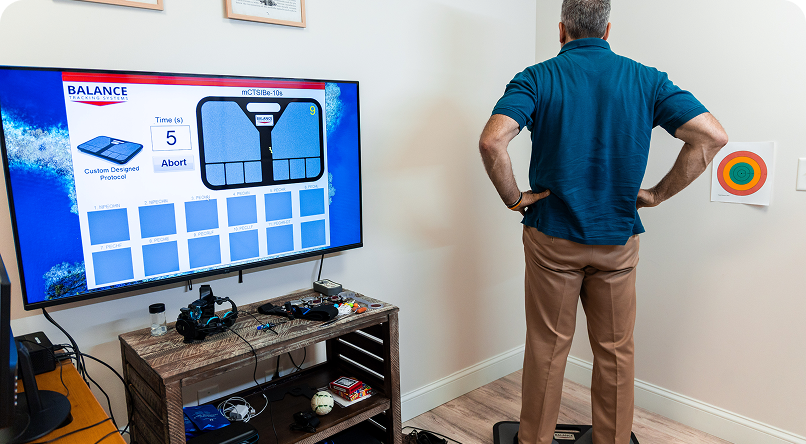 Man using a balance board, looking at a TV screen displaying a balance test in a room.
