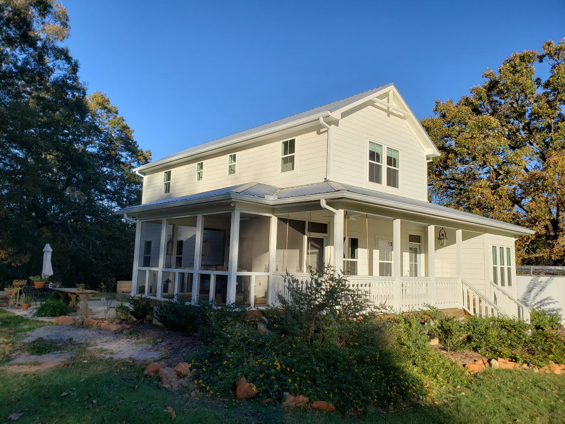White two-story house with porch under a clear blue sky. Bushes and trees surround the home.
