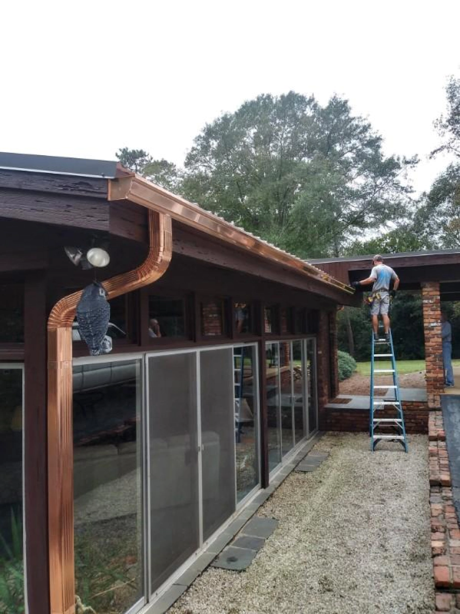 A person on a ladder installs copper gutters on a low-rise brown building with large windows.