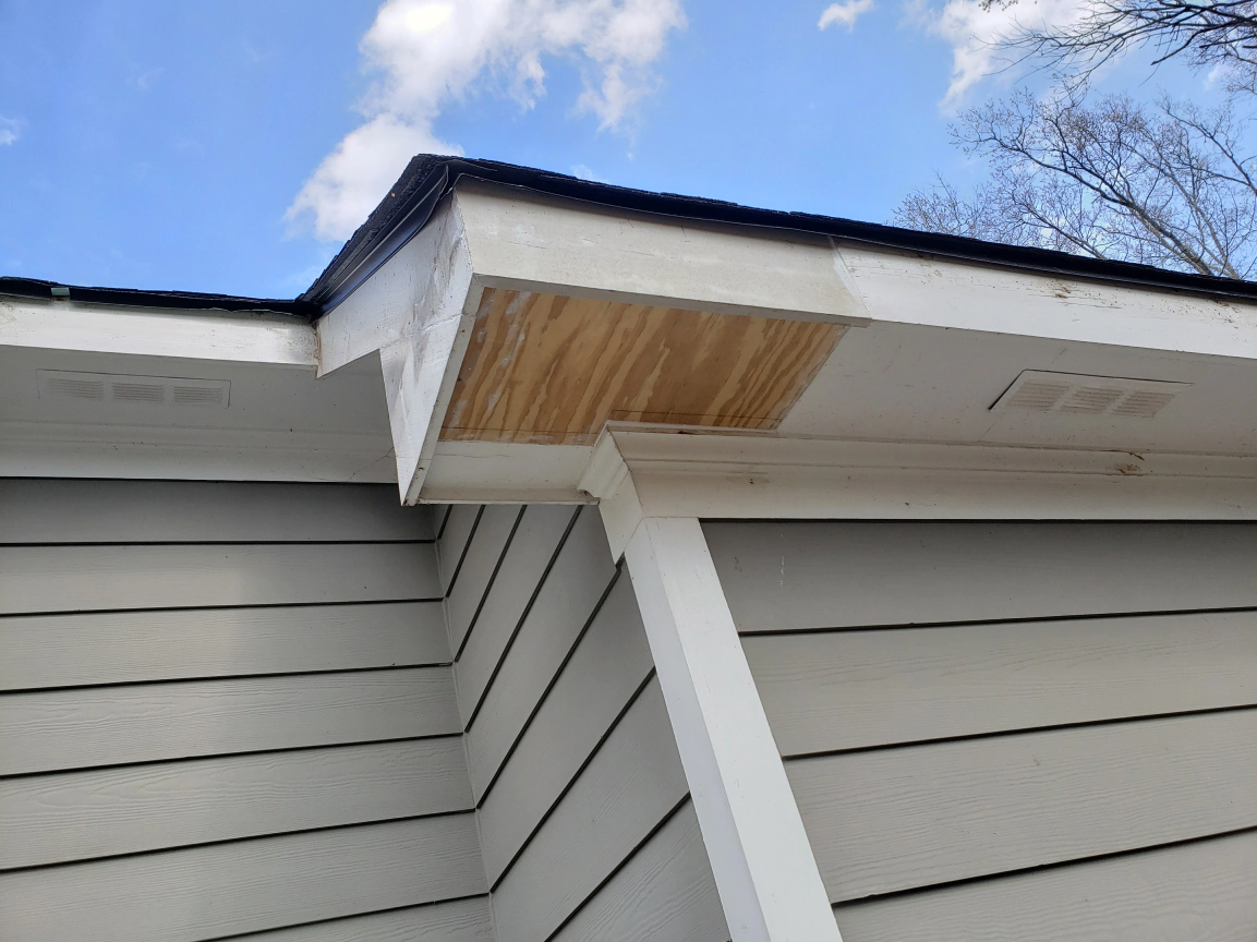 Exterior corner of a building, showing wood rot damage to the soffit, with blue sky above.