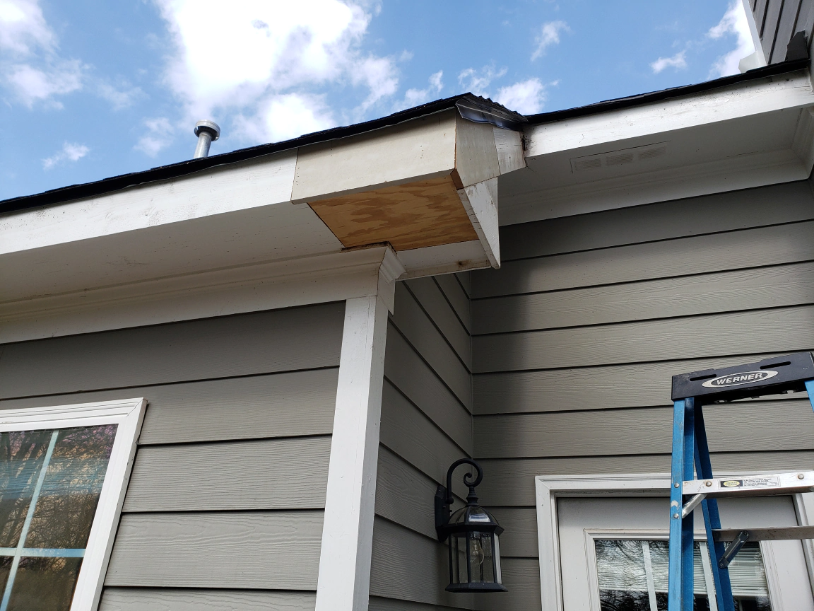 Exterior view of a house with visible soffit damage. A wooden panel is exposed on the corner.
