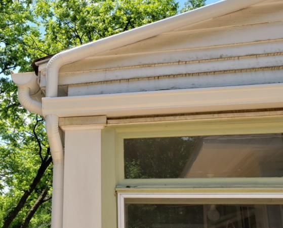 White gutters and siding of a house with visible dirt, window, and downspout.