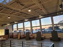 Interior of a building with large windows, couches, and an exposed ceiling, overlooking a city at sunset.