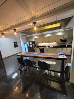 Spacious office kitchen with a long wooden table, chairs, and cabinets. Black floors and appliances.