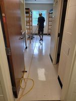 Person cleaning a room with a long cord. Room has open door, shelves, and a window.
