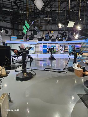 News studio with cameras, lights, and set; gray floor, blue and white background.