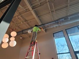 Person on a ladder cleaning ventilation ducts in an open room with large windows and hanging globe lights.