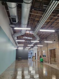 Interior view of a commercial space with exposed ductwork, concrete floor, and pendant lights; a worker cleans the floor.