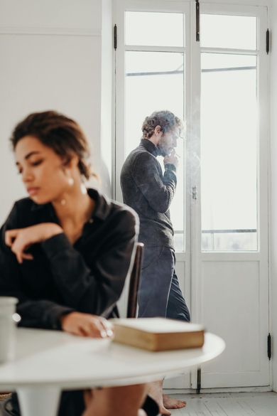 Uma mulher está sentada à mesa com um livro e um homem está de pé ao fundo.