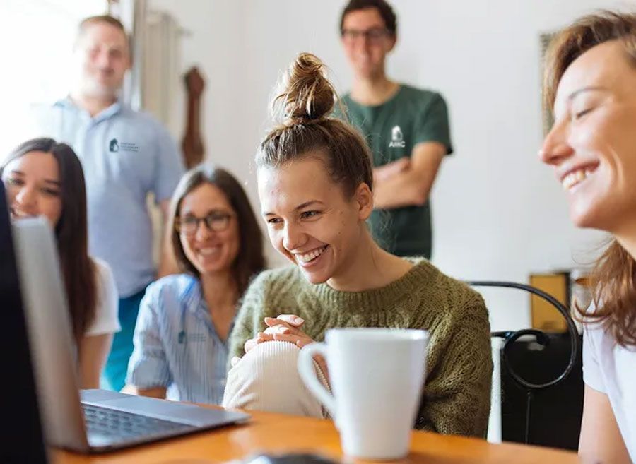 A group of friends smiles and laughs while looking at a laptop screen in a bright indoor setting.