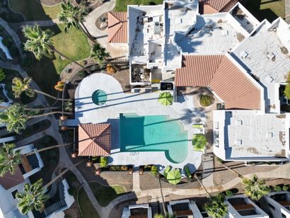 Aerial view of a resort with a pool, lounge chairs, palm trees, and buildings with terra-cotta roofs.