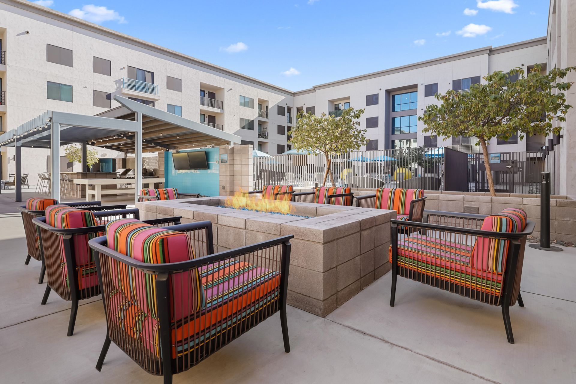 Outdoor lounge area with fire pit, chairs with colorful patterned cushions, and buildings in the background.