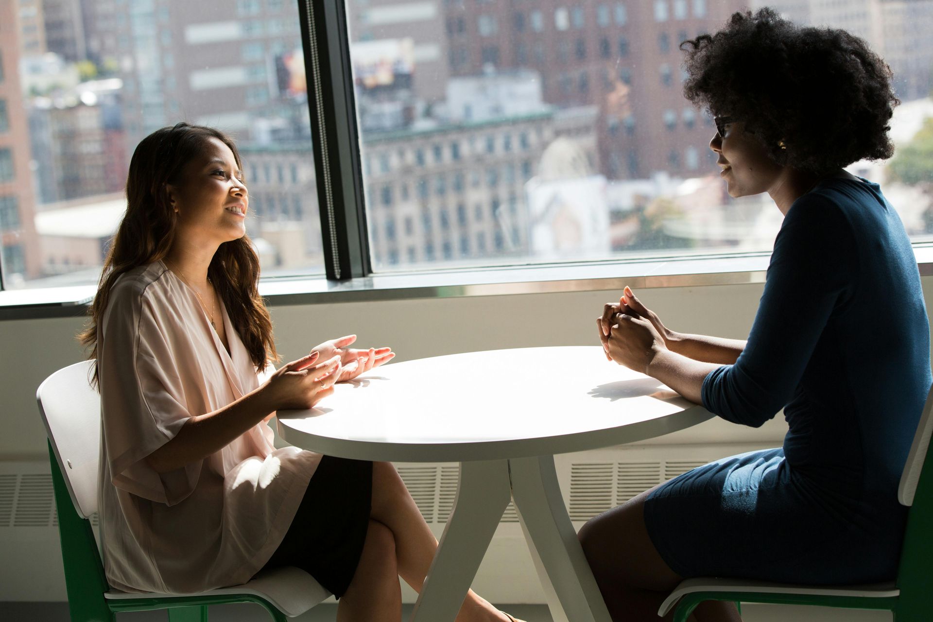 Two women talking at a round table near a window with city views. One gestures, the other listens.