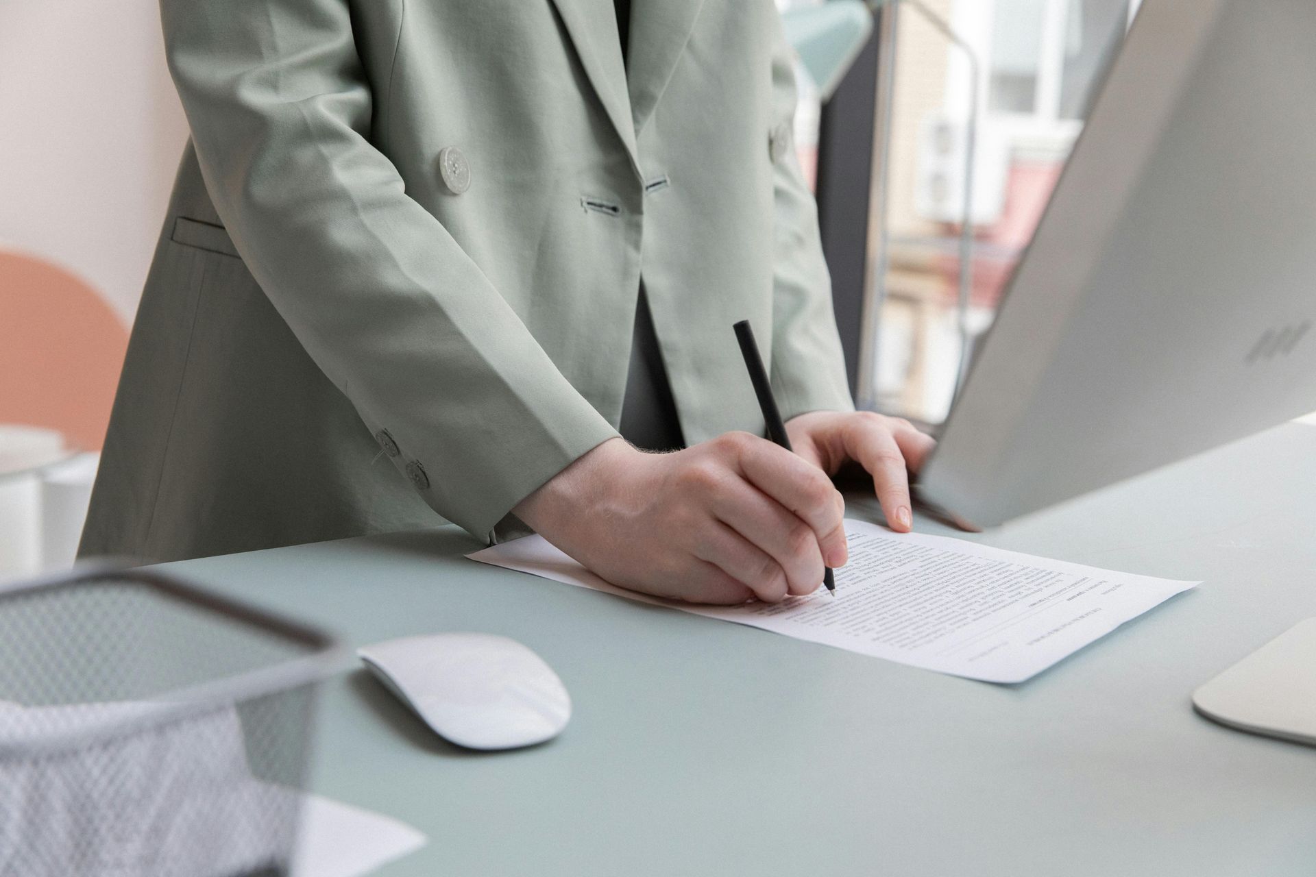 A person in a light green blazer writes on a paper at a desk with a computer and a mouse.