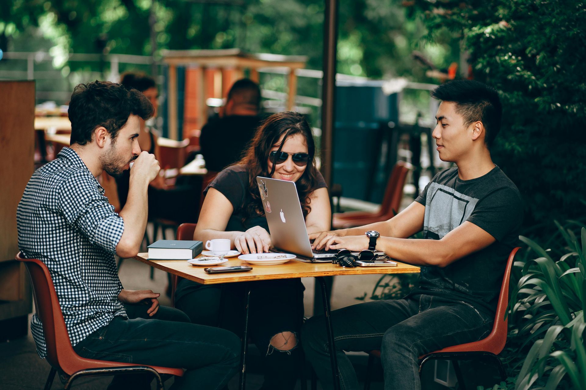 Three people sitting at a small table outdoors, working on a laptop. Two men and a woman are in conversation.