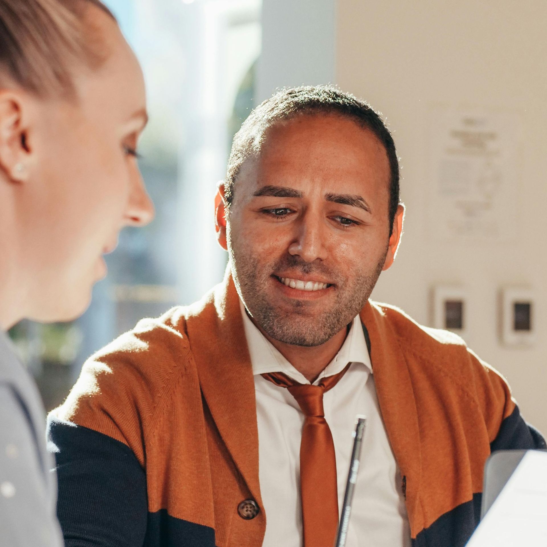 Man in a brown cardigan and tie smiles while looking at something with a woman; office setting.