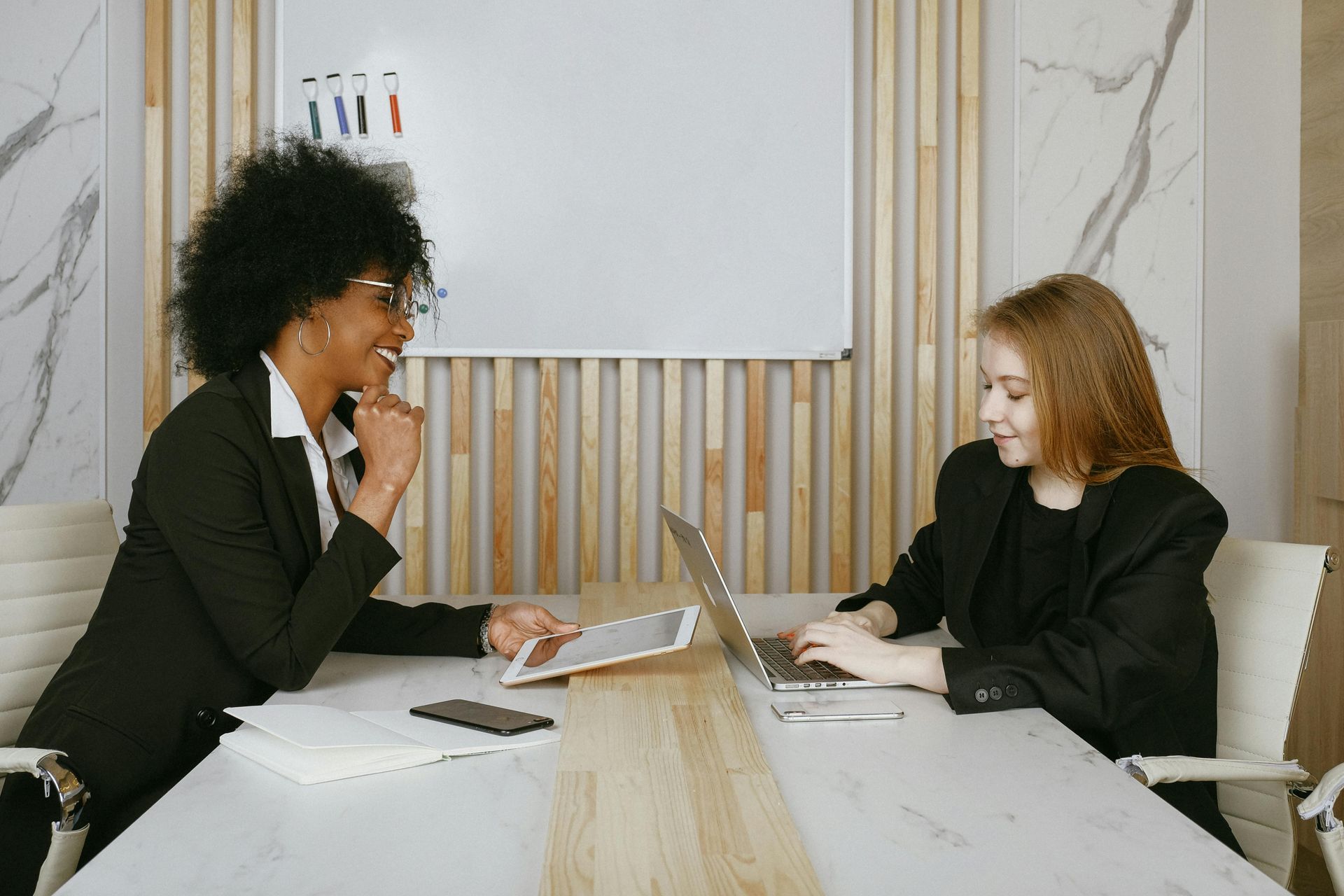 Two women in suits at a table, one with tablet, other on laptop, smiling in a modern office.