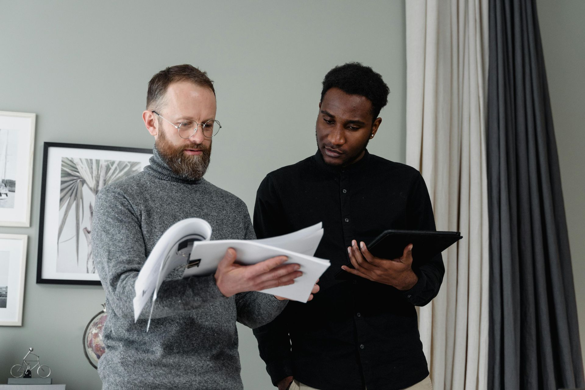 Two men in room reviewing documents, one with tablet.