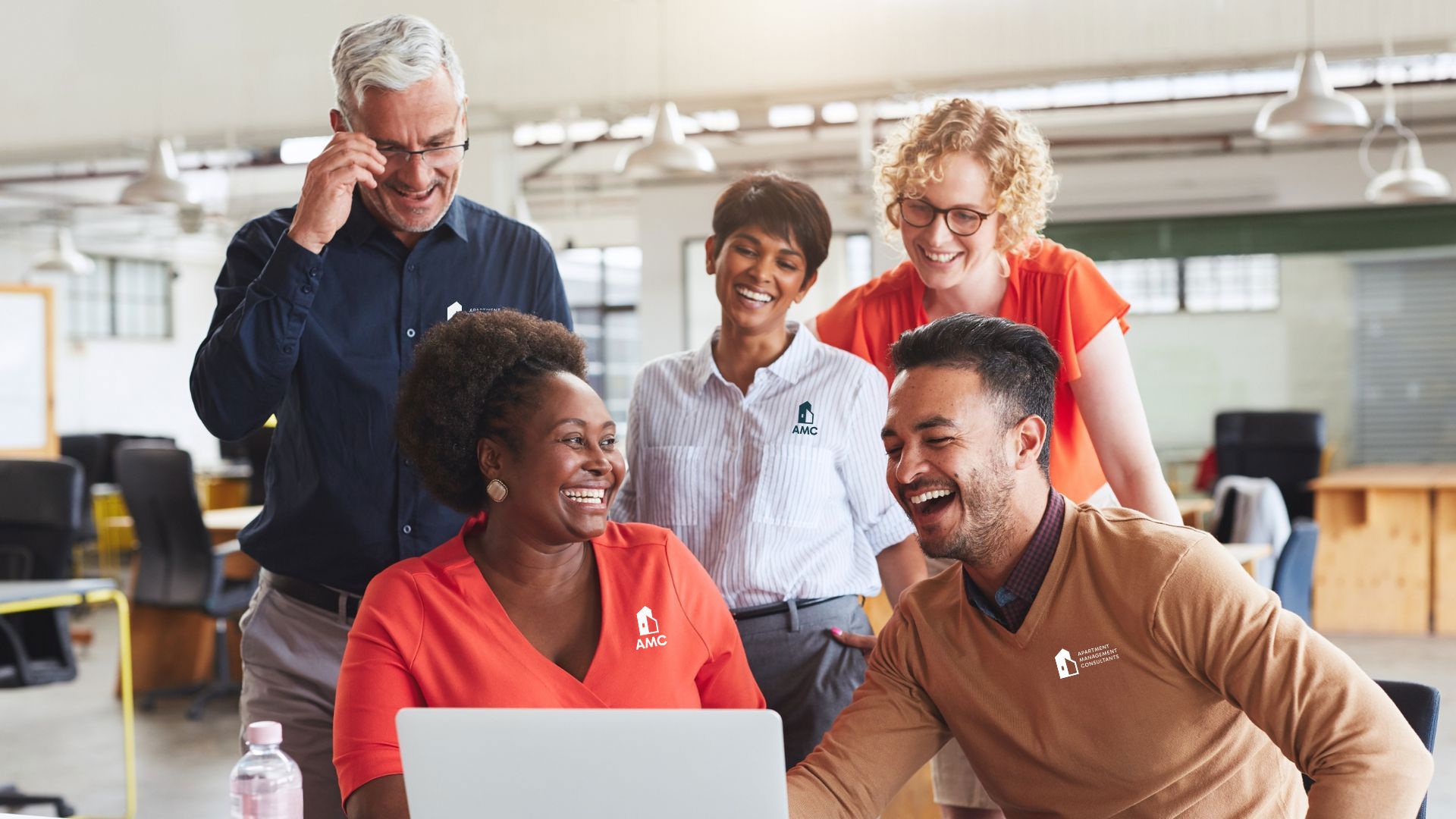 Group of diverse colleagues smiling and laughing while looking at a laptop in a bright office setting.