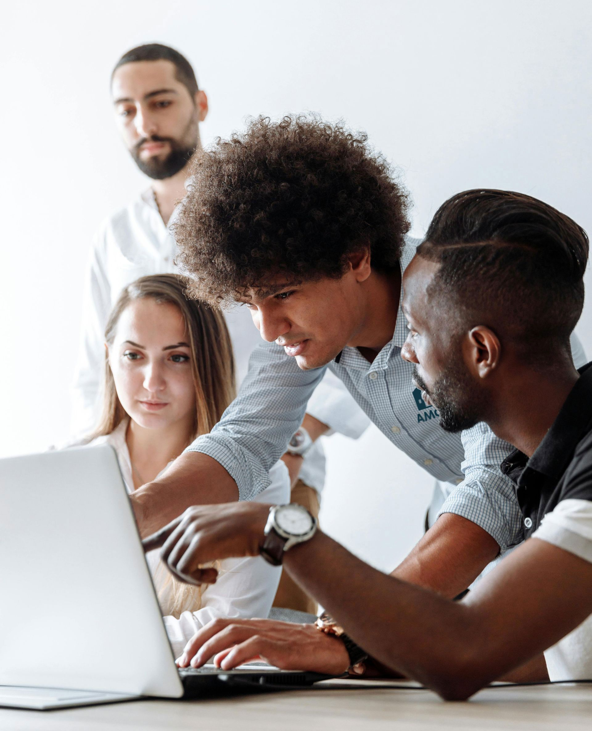 Four diverse colleagues collaborate at a laptop, pointing and discussing. They are indoors, possibly in an office setting.