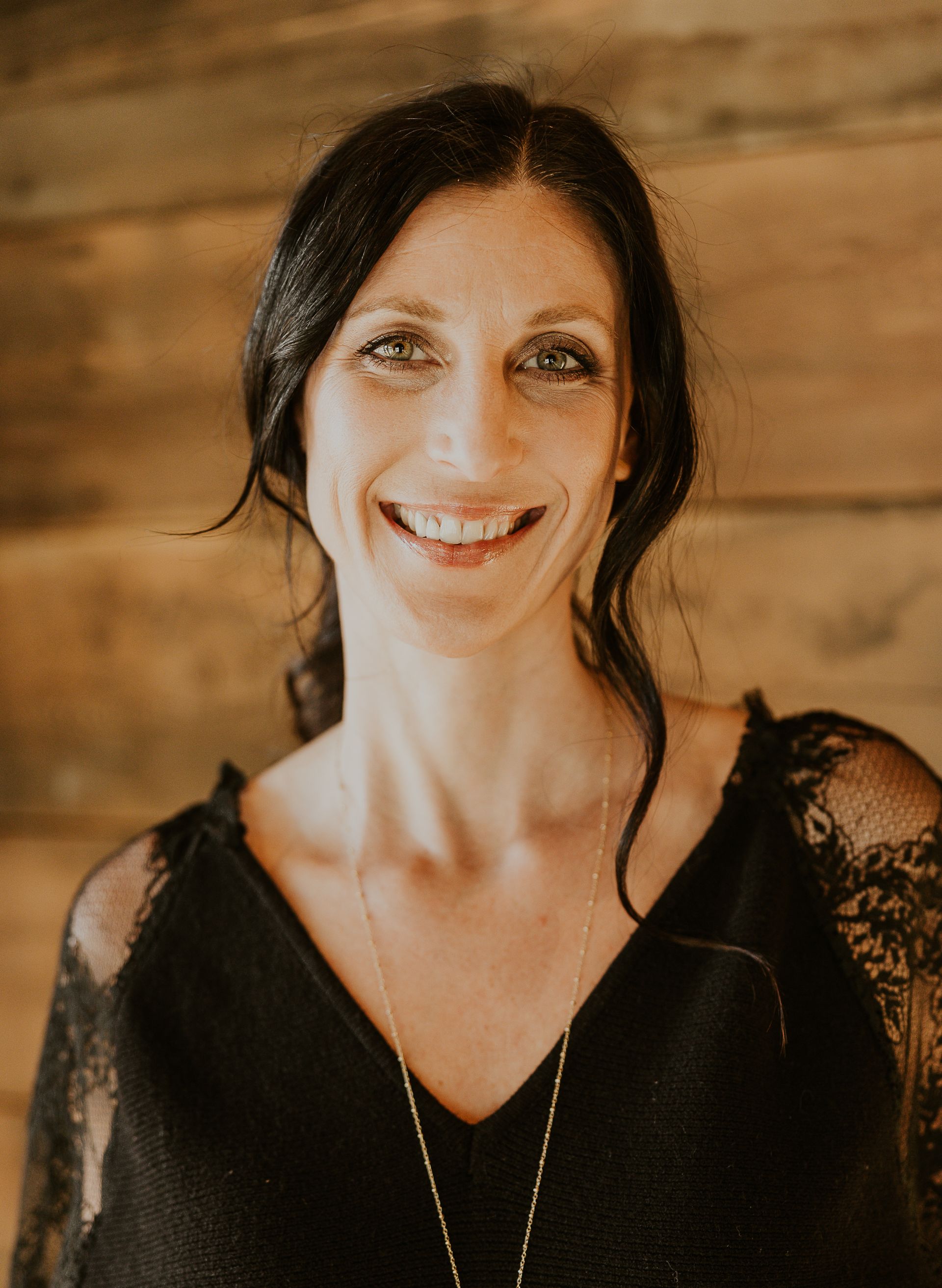 A woman in a black dress is smiling in front of a wooden wall.