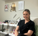 Beauty therapist smiling in a modern home beauty studio surrounded by skincare and wellness products, treatment tools, and calming décor.