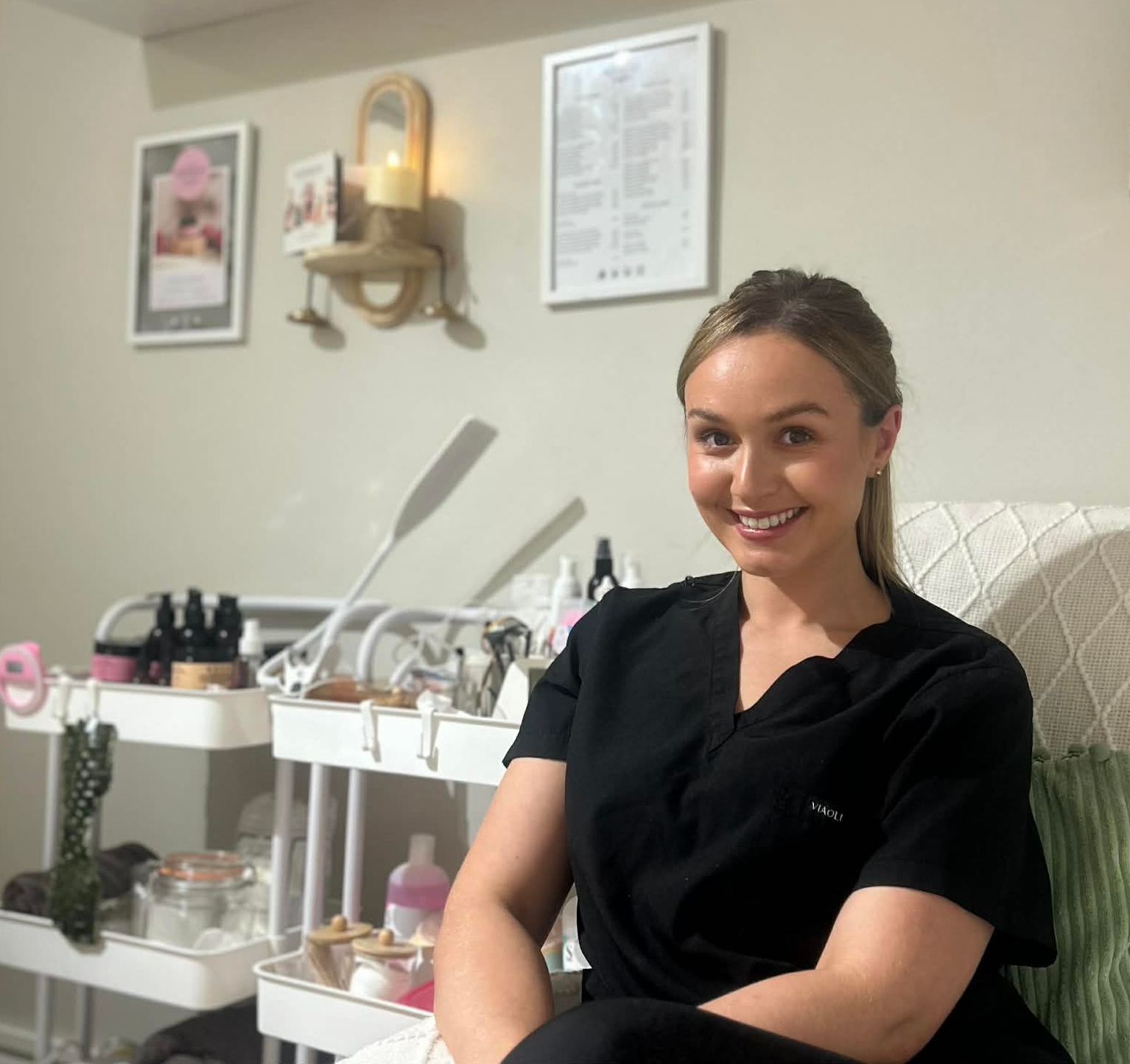 Beauty therapist smiling in a modern home beauty studio surrounded by skincare and wellness products, treatment tools, and calming décor.