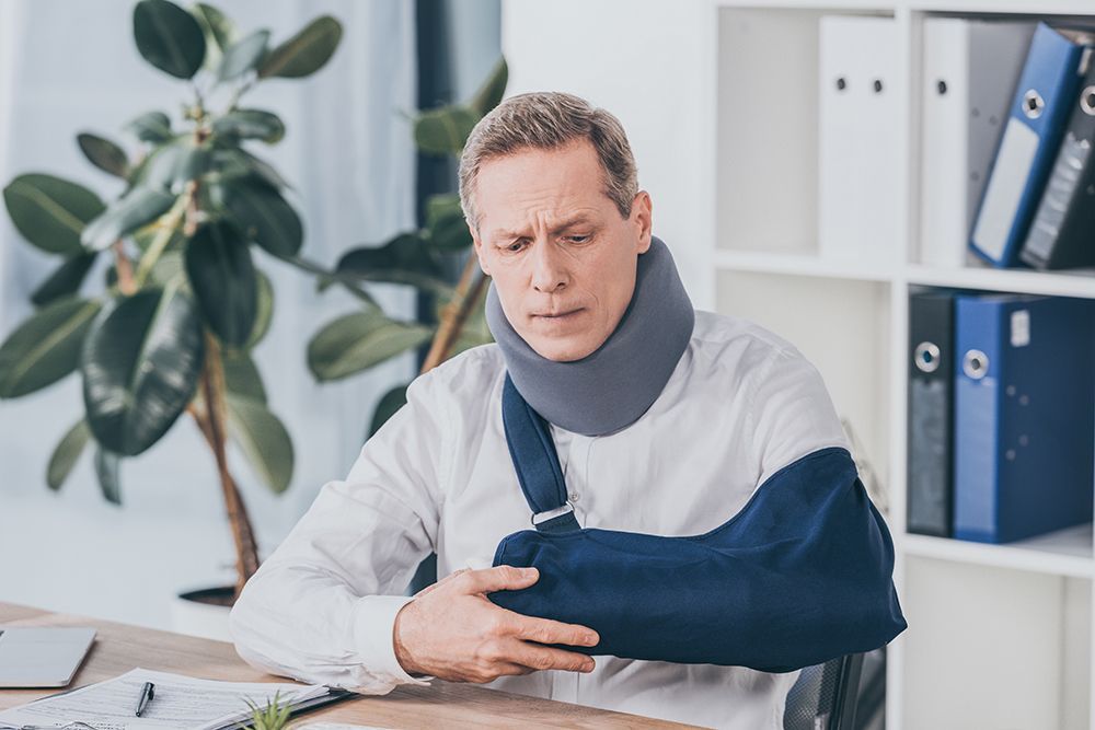 middle aged worker holding broken arm wile sitting at table with document in office, compensation concept