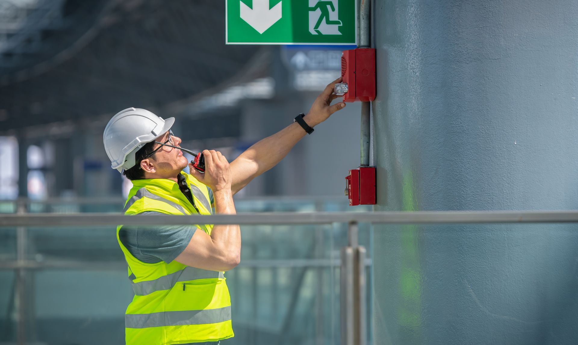 Worker in safety vest testing fire alarm under emergency exit sign indoors.