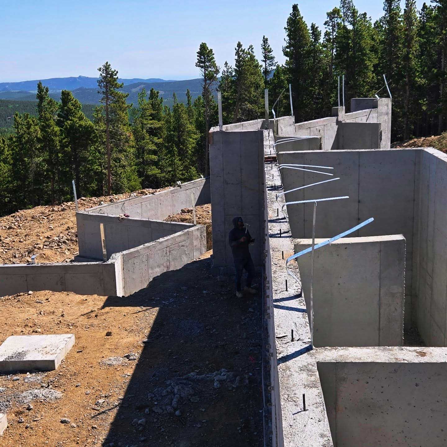 Concrete foundation under construction on a hillside. A worker stands near the wall. Background includes trees and a mountain view.