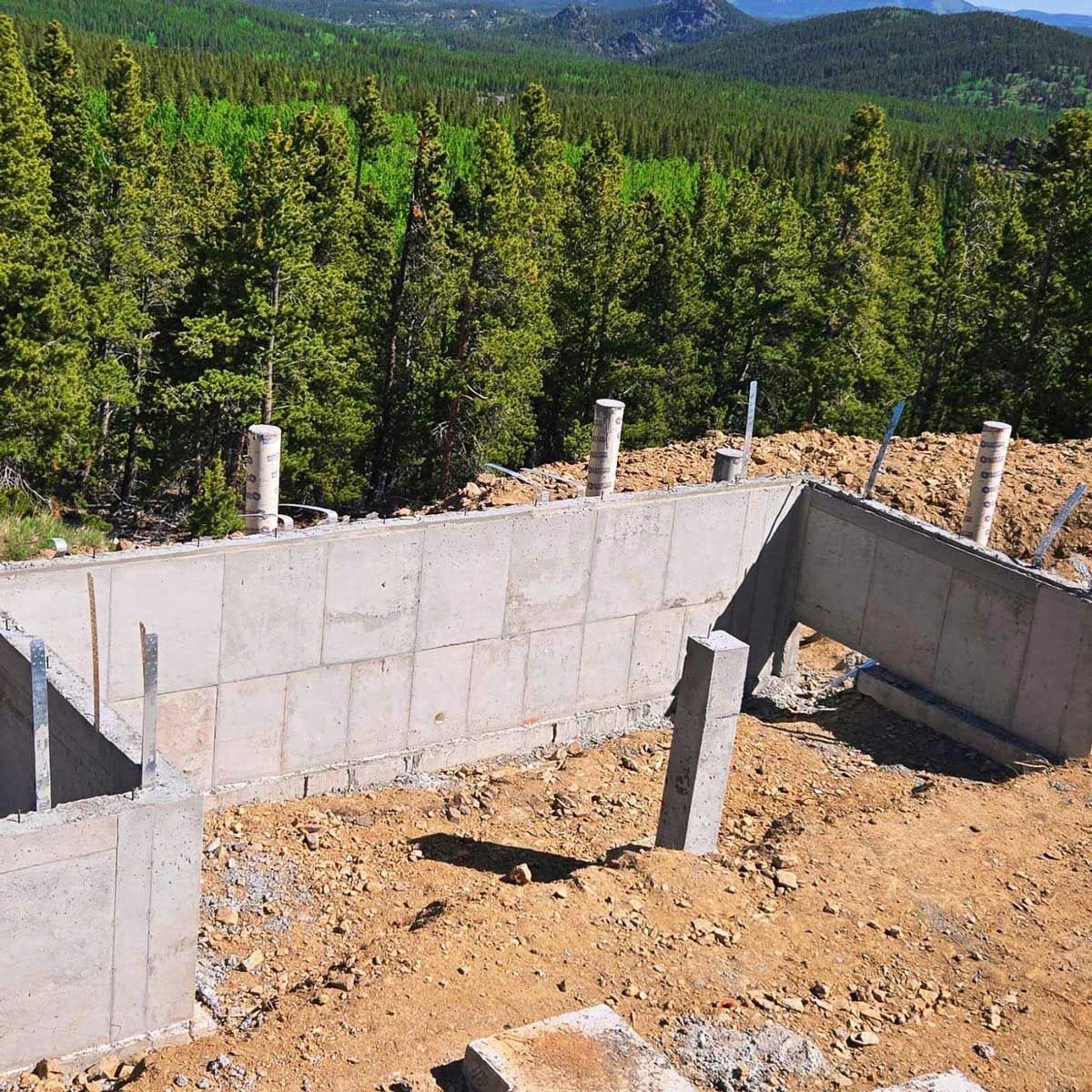 Concrete foundation of a house under construction against a backdrop of a green forest and mountains.