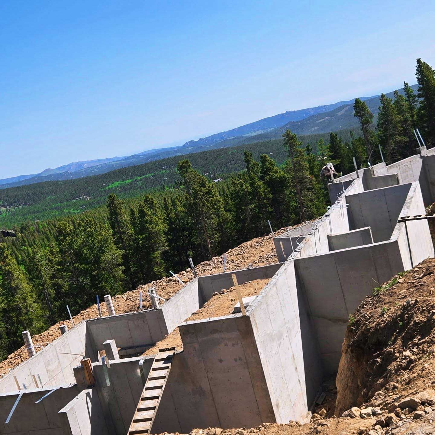 Concrete foundation being built on a sloped hillside with a mountainous background and trees under a blue sky.