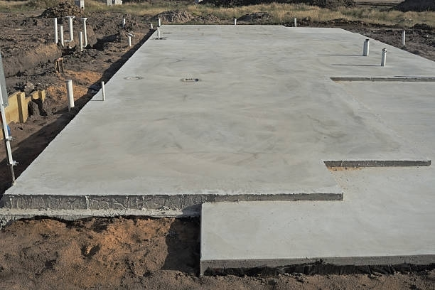 A freshly poured concrete foundation for a building, with pipes and trenches visible in an outdoor setting.