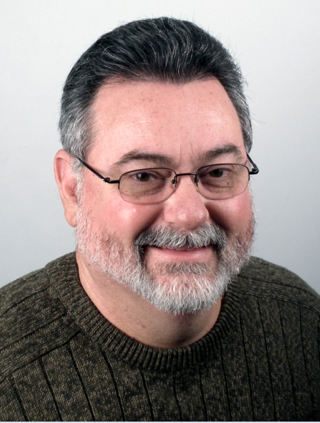 A person with short, light brown hair wearing dark-rimmed glasses and a navy blue floral patterned top against a gray wall.