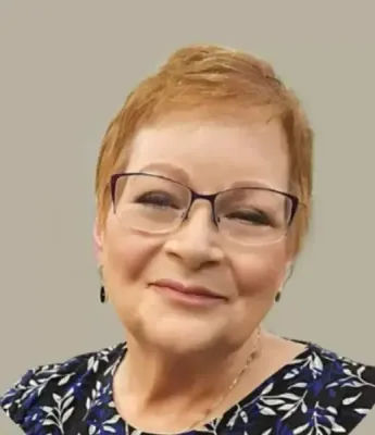 A person with short, light brown hair wearing dark-rimmed glasses and a navy blue floral patterned top against a gray wall.