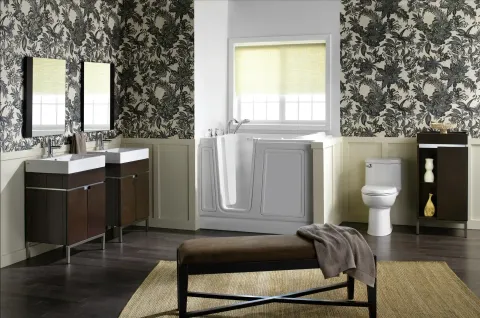 A modern bathroom with dual vanities, floral wallpaper, a walk-in bathtub, and a toilet with a bench in the foreground.