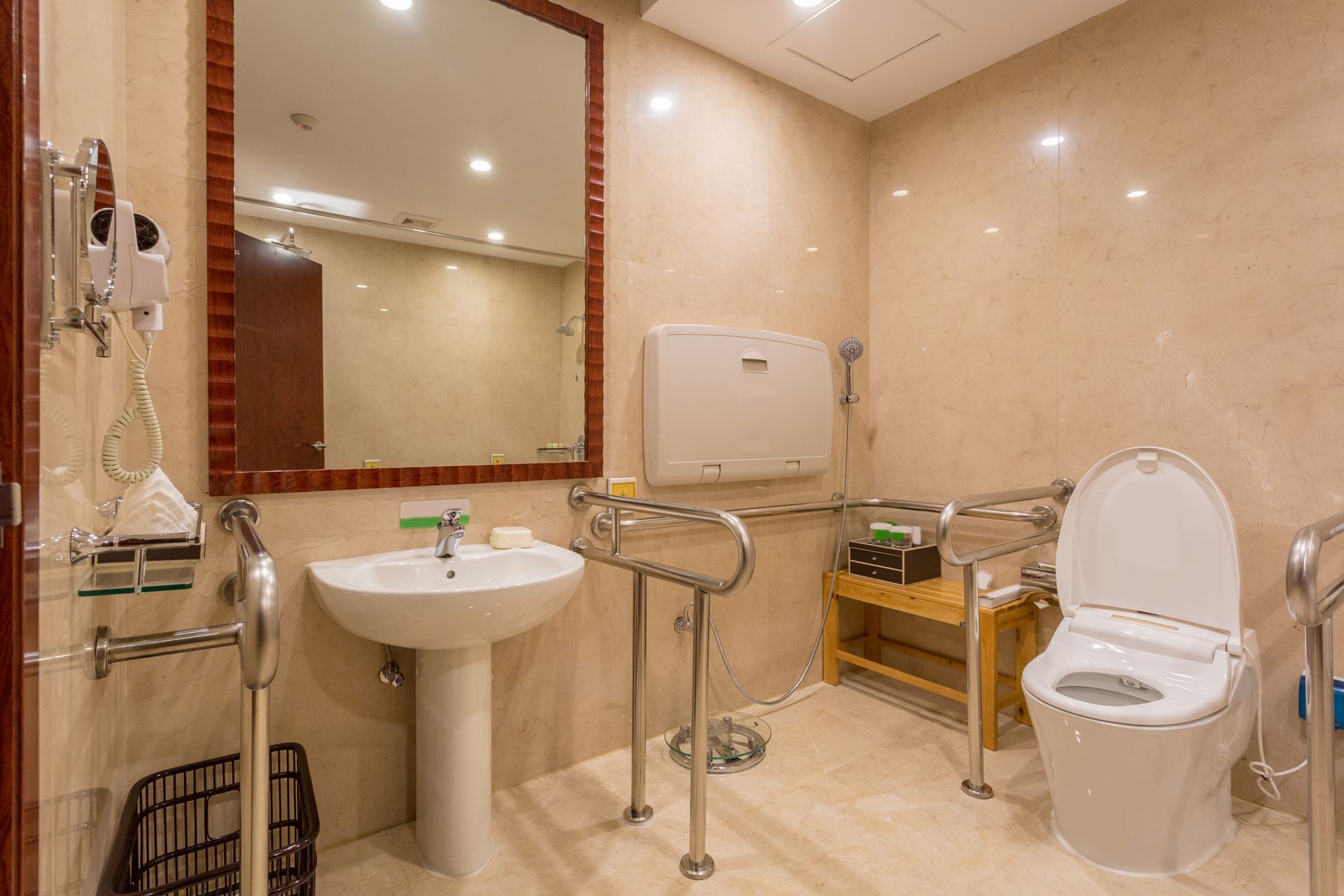 Accessible bathroom with sink, mirror, grab bars, toilet, and shower seat in beige tiles