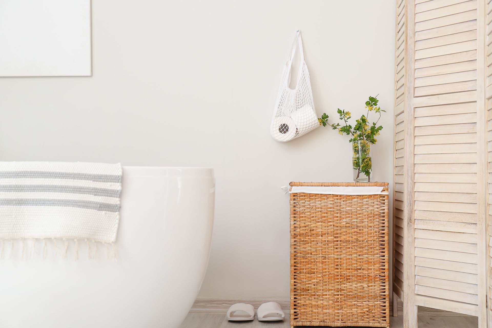 Minimal bathroom corner with white tub, wicker basket, plants, and slippers near a louvered door