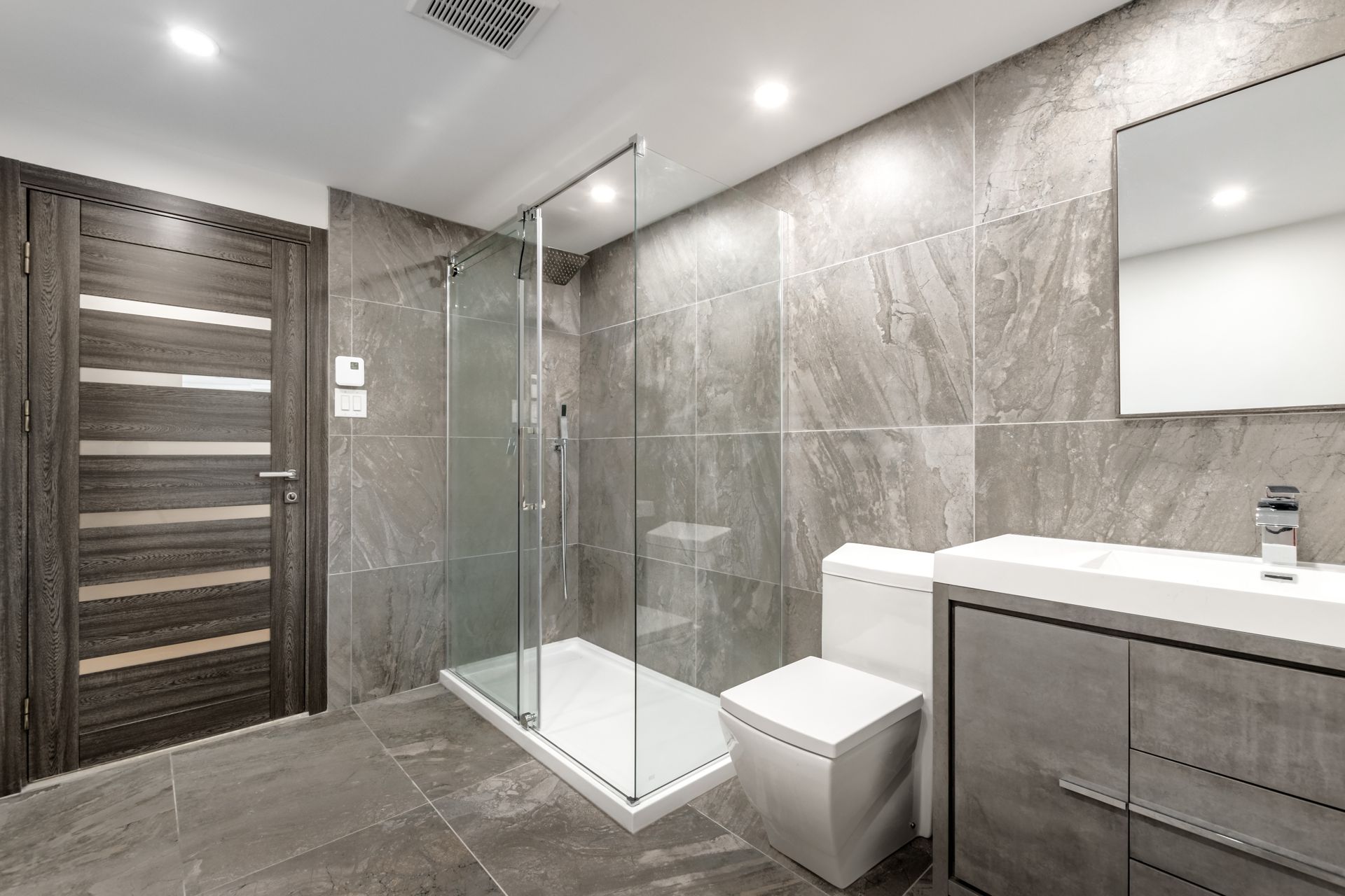 Modern gray bathroom with glass shower, vanity, toilet, and wood door