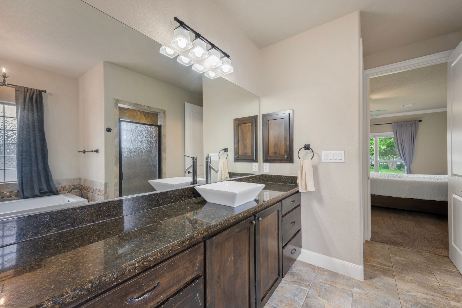 Modern bathroom with double vanity, granite countertop, mirrors, and a bedroom visible through an open doorway