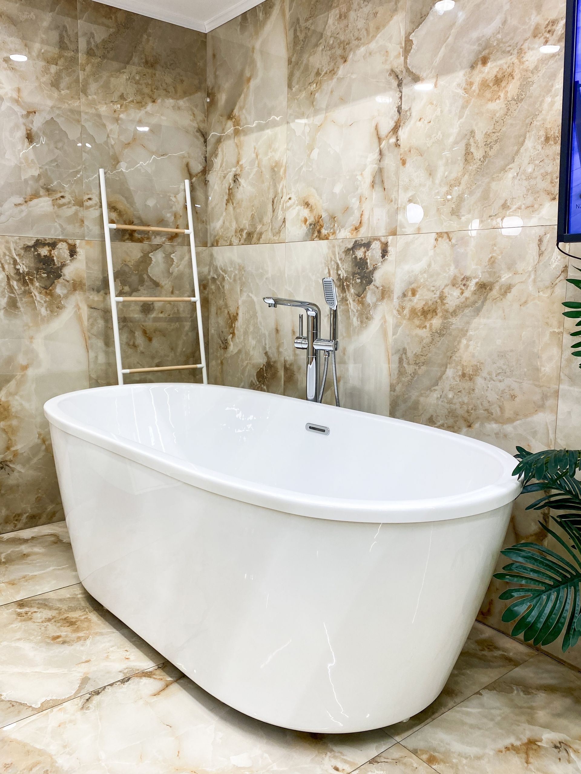 White freestanding bathtub in a beige marble-tiled bathroom with chrome fixtures and a ladder towel rack