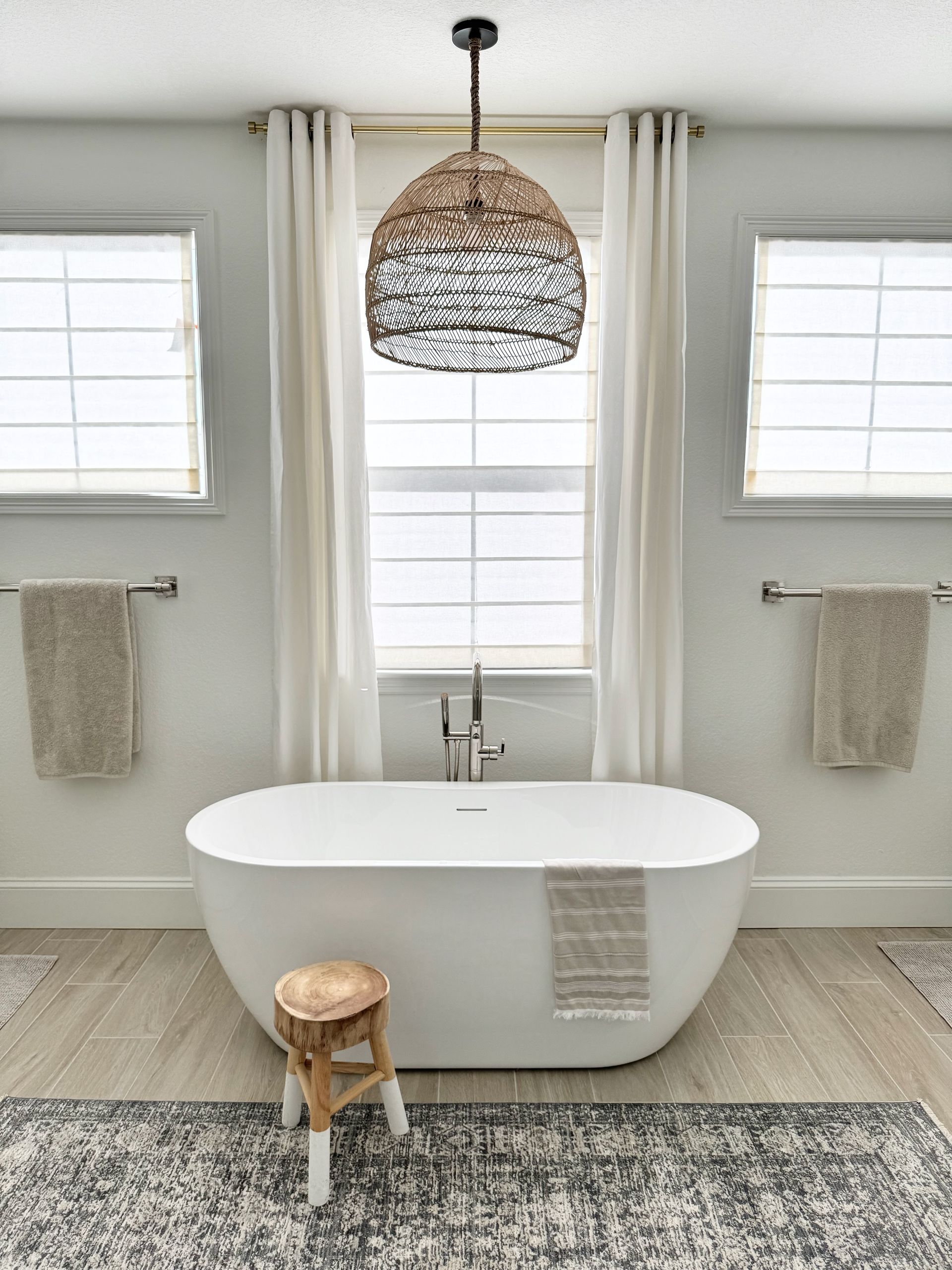 Bright bathroom with freestanding tub, woven pendant light, and neutral decor, centered between two windows.