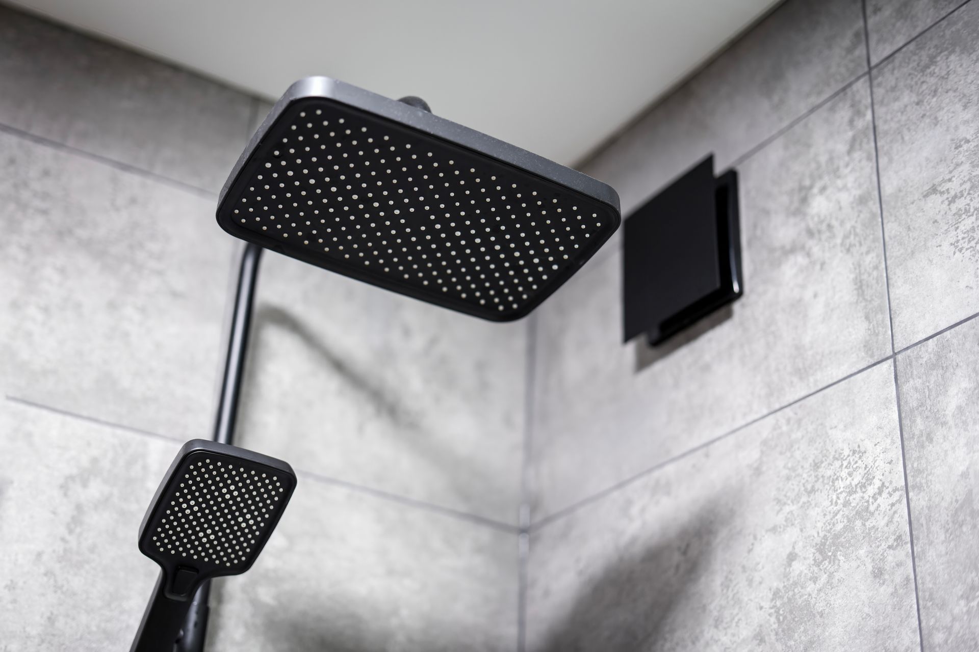 Modern black shower heads in a gray-tiled bathroom, with one large overhead and one handheld showerhead.