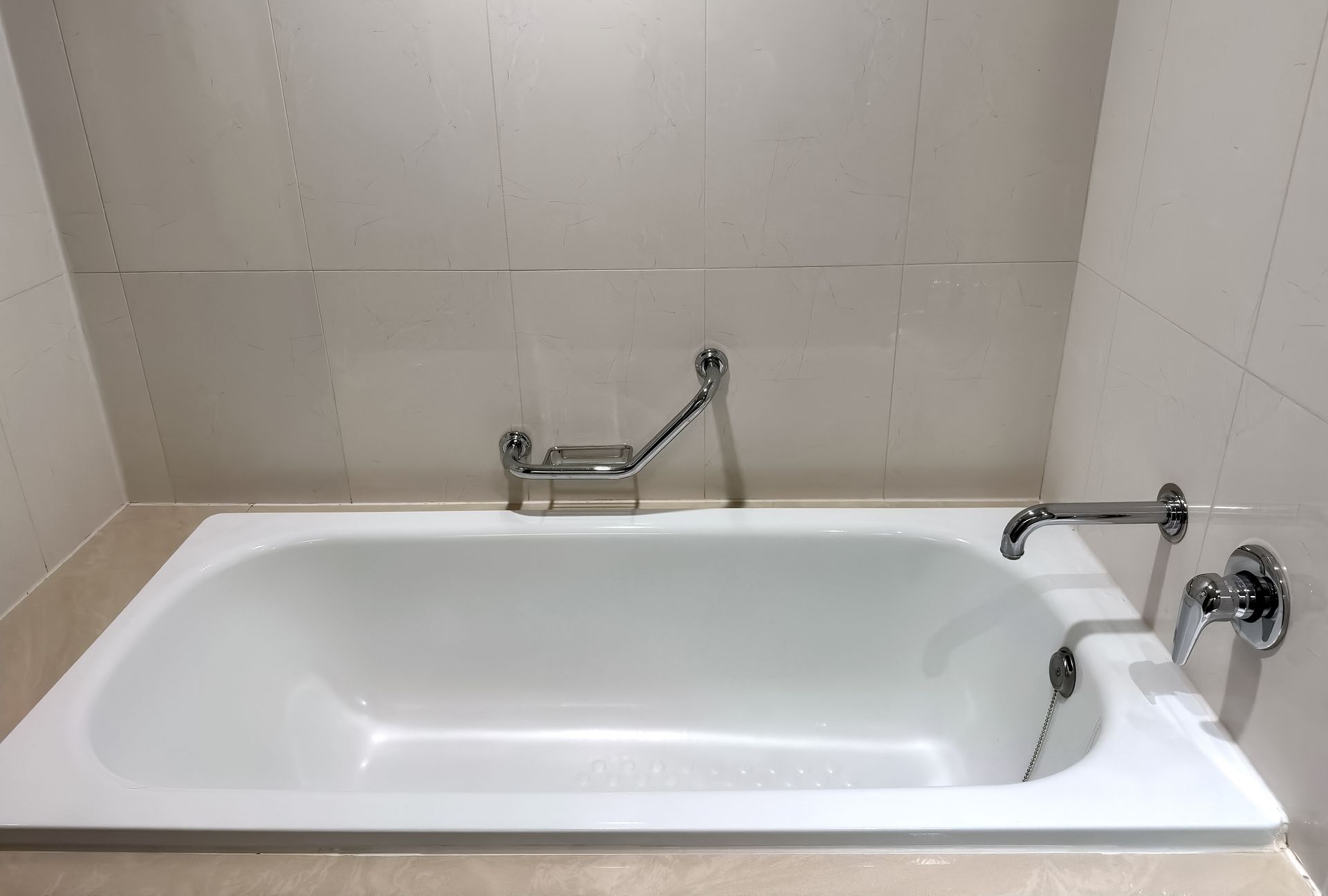 Empty white bathtub with chrome faucets against beige tiled bathroom walls