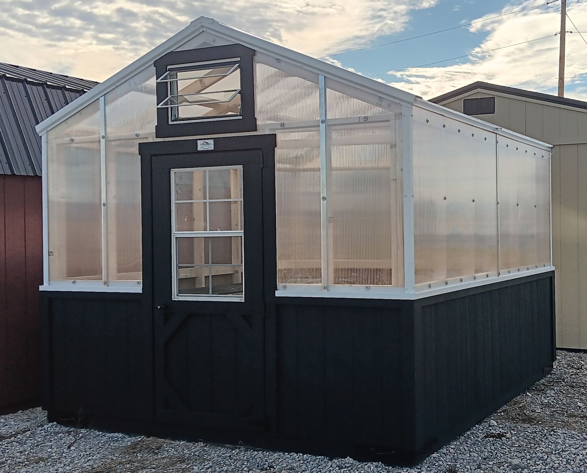Greenhouse with black base and clear, corrugated plastic walls, white trim, and a single door. Situated outdoors on gravel.