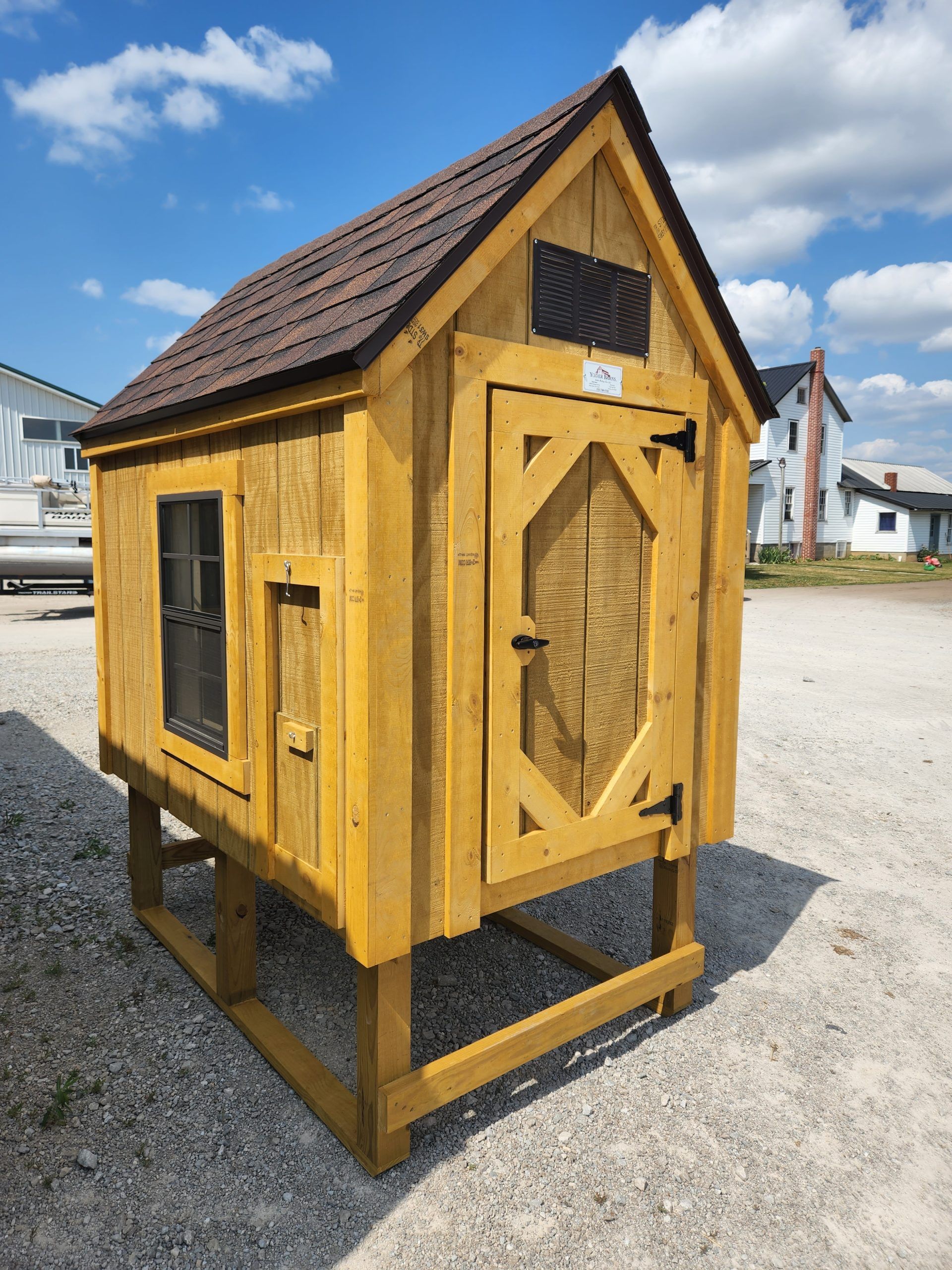 Yellow wooden chicken coop with a dark roof, standing on a gravel surface under a blue sky.
