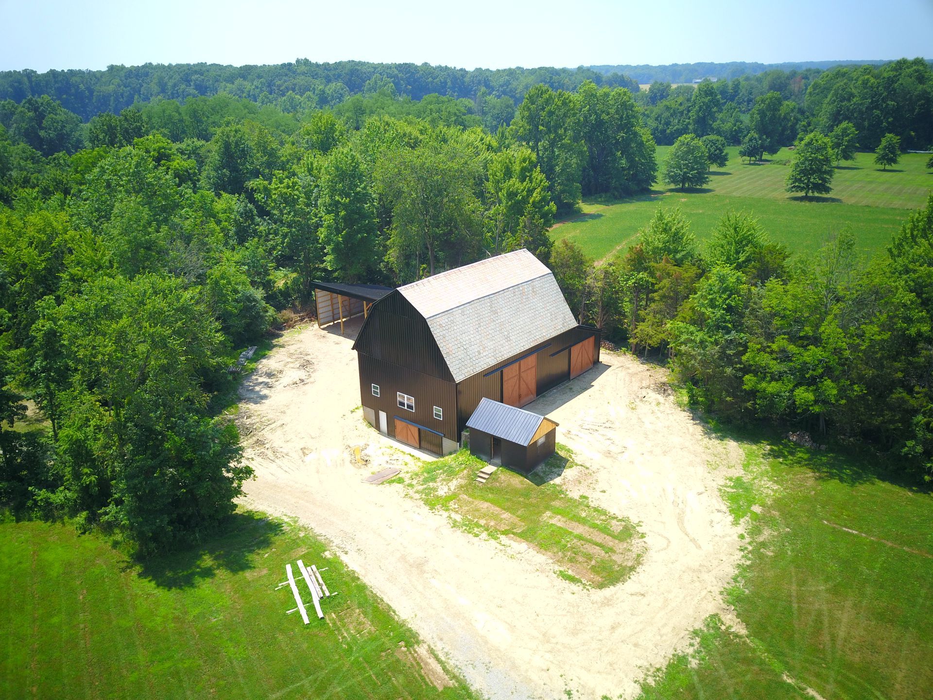 Barn in a clearing surrounded by green trees and grass, under a blue sky. A gravel driveway leads up to it.